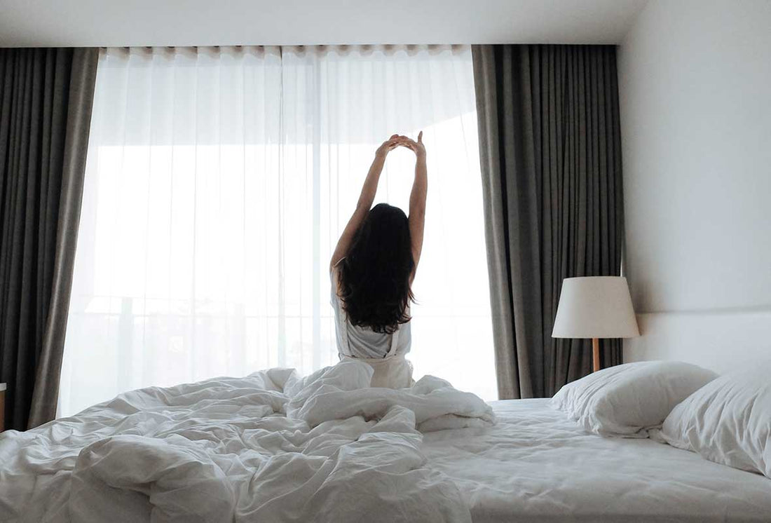 A woman sitting on an unmade bed stretching in front of a window