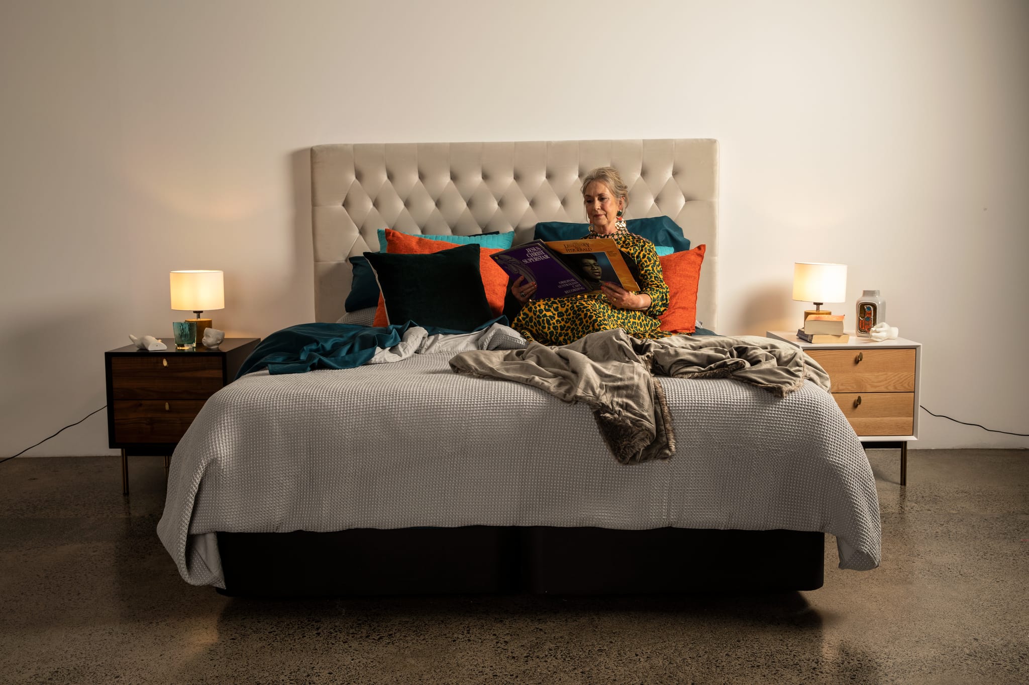A woman sits on a SleepMaker bed with a light beige tufted headboard, surrounded by colorful pillows, a gray comforter, and rumpled blankets in a modern bedroom setting.
