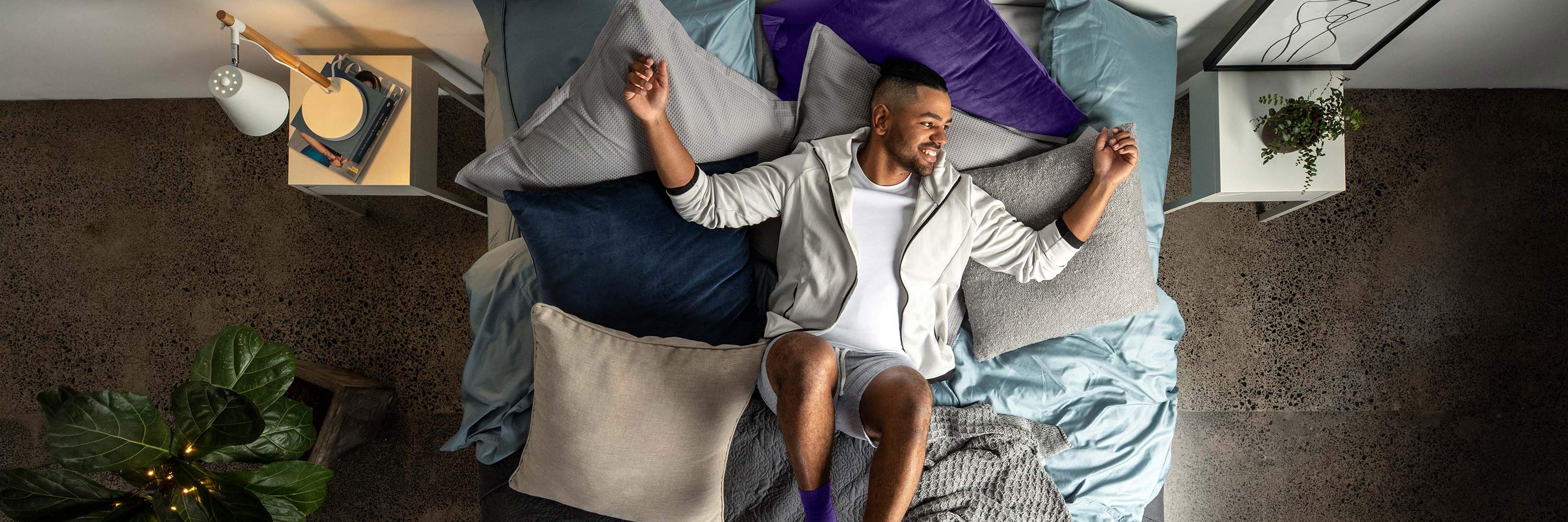 Man lying on a SleepMaker bed surrounded by various pillows in a bedroom setting.