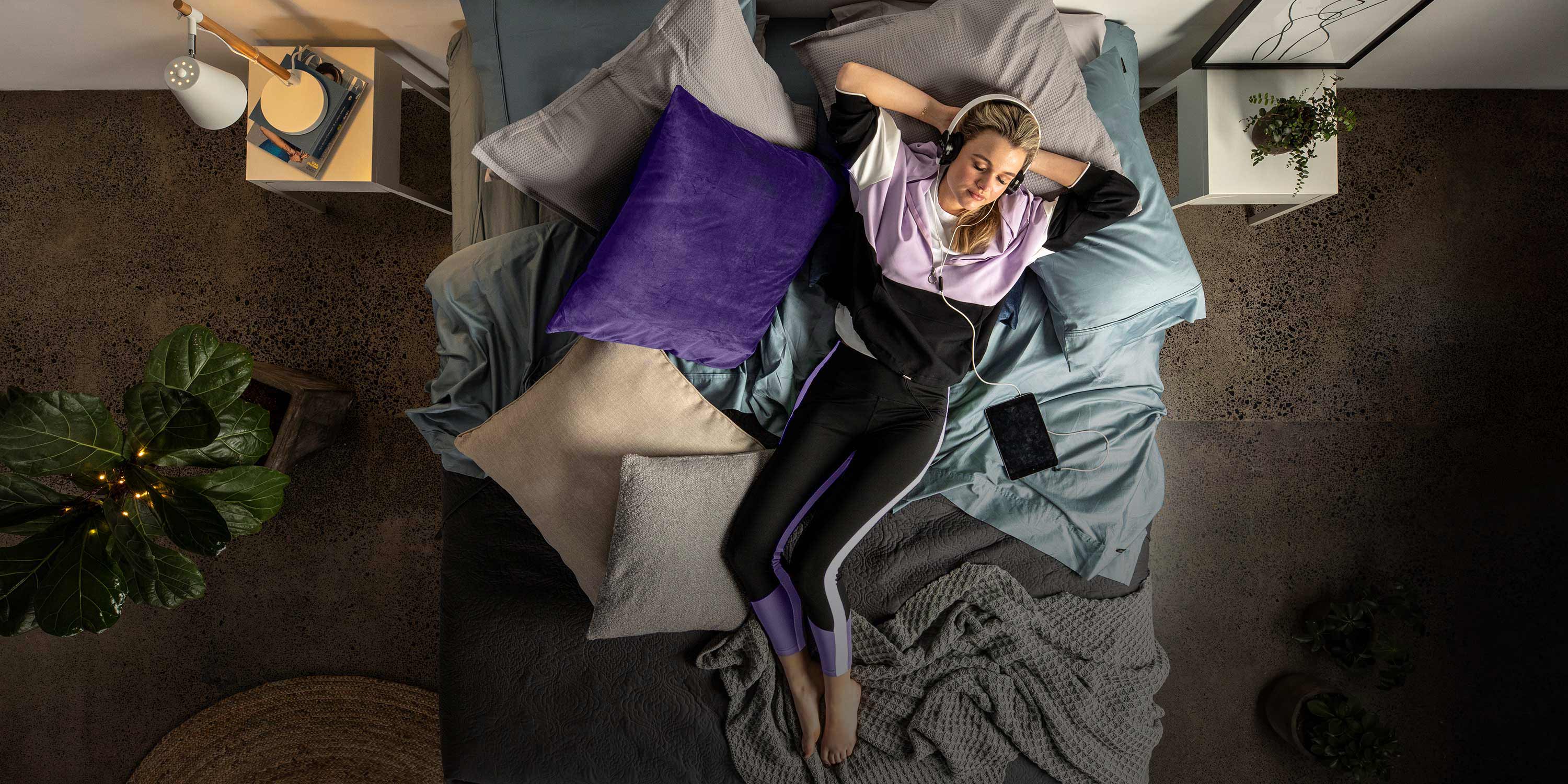 Young woman lying on a bed with headphones on, surrounded by pillows and a plant.